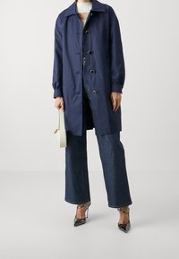 Granatowy wodoodporny płaszcz z klasycznym kołnierzem, zapinany z przodu na guziki, z elastycznymi mankietami, w połączeniu z rozszerzanymi dżinsami i metalicznymi wysokimi obcasami.