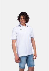 Polo bianco con colletto e polsini a strisce nere e blu, con un piccolo logo sul lato sinistro del petto. Indossato con pantaloni corti in denim azzurro.