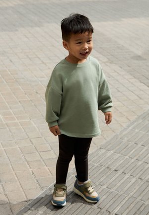 Ung dreng står på fortovet iført en grøn oversized sweatshirt, sorte bukser og sneakers, smilende og kigger til siden.