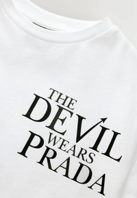 Chemise blanche avec texte noir "THE DEVIL WEARS PRADA", avec une petite queue de diable intégrée dans la lettre "I" du mot "DEVIL".