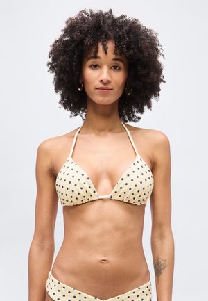 Žena s kučeravými vlasmi v béžových bikinách s námorníckymi modrými bodkami, stojaca pred jednoduchým svetlým pozadím.