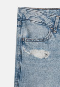 Jean en denim bleu clair présentant un design déchiré avec un grand trou et des bords effilochés. Couture visible en marron contrasté.