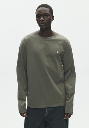 Ung person iført en olivengrøn langærmet skjorte med et lille rundt logo på venstre bryst, kombineret med mørkeblå jeans.