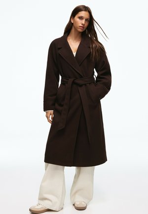 Femme portant un long manteau marron foncé ceinturé, un pantalon large blanc et des baskets beiges, debout devant un fond clair uni.