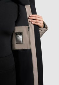 Interior de chaqueta negra y beige con un bolsillo con cremallera y etiqueta de cuidado. Textura suave con paneles de color contrastante; mano visible en el borde.