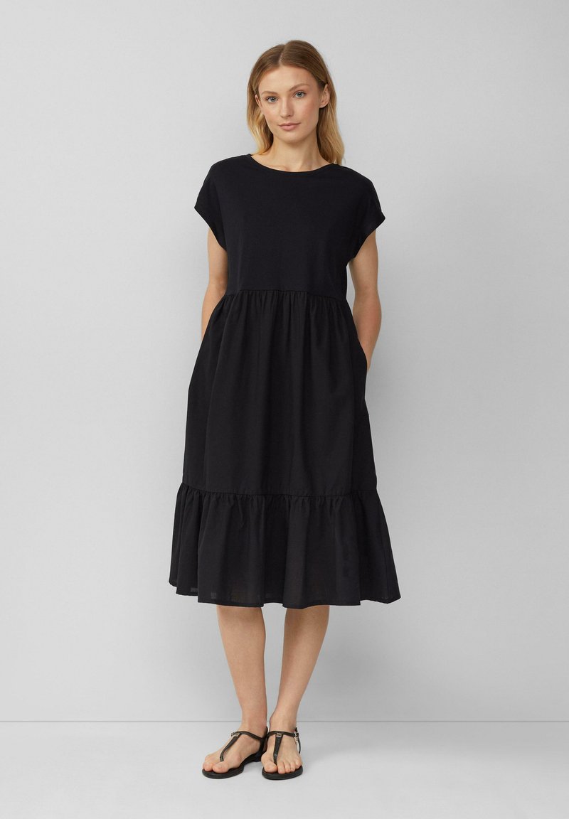 Schwarzes Baumwollkleid mit kurzen Ärmeln, rundem Ausschnitt und schwingendem, gestuftem Rock. Verfügt über seitliche Taschen und eine weiche, leichte Textur.
