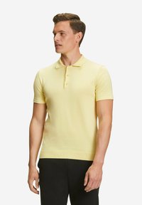 Polo in morbido tessuto giallo chiaro, con colletto classico, maniche corte, chiusura a tre bottoni e orlo a coste. Indossato con pantaloni scuri.