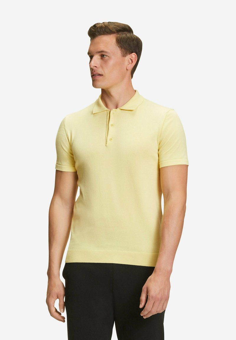 Polo in morbido tessuto giallo chiaro, con colletto classico, maniche corte, chiusura a tre bottoni e orlo a coste. Indossato con pantaloni scuri.