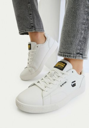 Zapatillas bajas blancas de G-Star combinadas con jeans grises enrollados sobre un fondo blanco.