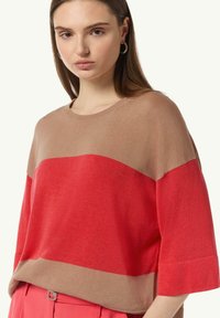 Sweter dzianinowy o wzorze kolorowych bloków w odcieniach beżu i jasnej czerwieni. Ma krótkie rękawy i luźny krój, a jego faktura jest gładka.