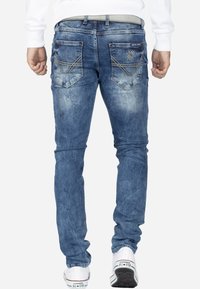 Jeans en denim bleu clair à coupe slim, avec des poches arrière ornées de coutures décoratives et de bords en angle. Associés à des baskets blanches.