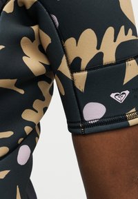 Κοντινό πλάνο του πάνω βραχίονα ενός ατόμου που φοράει μαύρο και καφέ ανάγλυφο κοντομάνικο καταδυτικό κοστούμι με ορατές ραφές και λογότυπο.