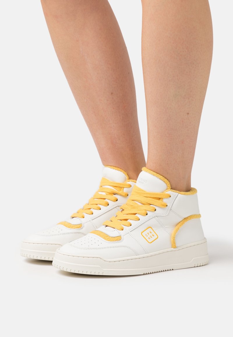 Baskets blanches montantes avec lacets et bordures jaunes portées sur des jambes nues sur un fond clair uni.