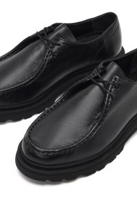 Zapatos de cuero negro con punta redonda, con cordones y detalles de costura, reposando sobre una suela de goma negra gruesa y texturizada.