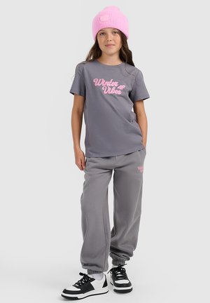Młoda dziewczyna ubrana w szary t-shirt z napisem "Winter Vibes", dopasowane szare spodnie dresowe, czarno-białe trampki i jasnoróżową dzianą czapkę.