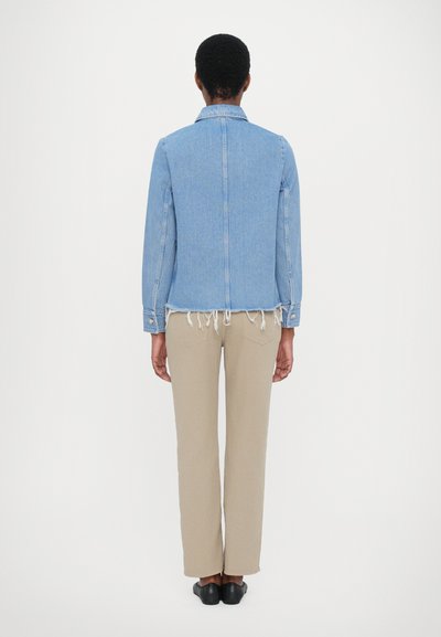 Denim jacket in light blue with a frayed hem, long sleeves, and snap buttons. Paired with beige straight-leg trousers and black shoes.