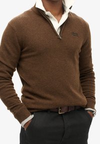 Uomo che indossa un maglione a collo zip bruno sopra una camicia bianca con colletto, pantaloni neri, con le mani nelle tasche e un orologio al polso sinistro.