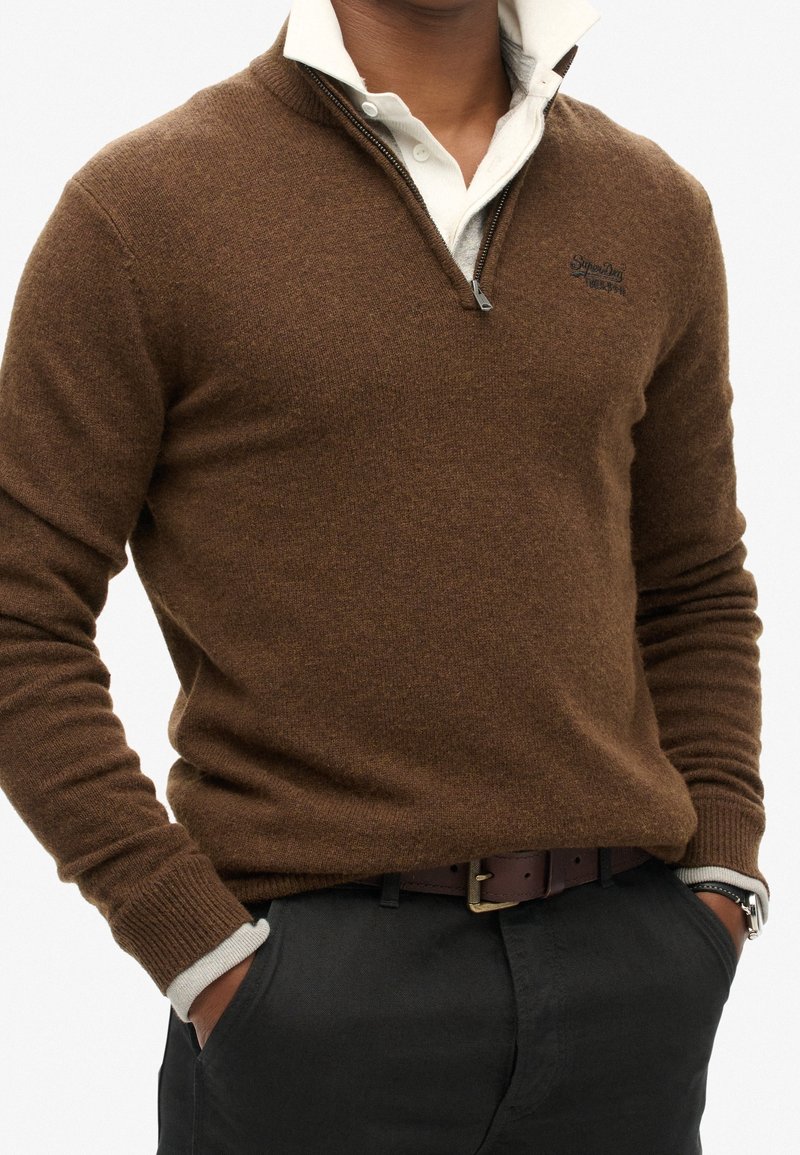 Uomo che indossa un maglione a collo zip bruno sopra una camicia bianca con colletto, pantaloni neri, con le mani nelle tasche e un orologio al polso sinistro.