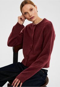Burgunderfärgad stickad cardigan med rund halsringning, knappstängning, ribbade muddar och en avskuren nederdel. Bärs över mörka jeans.