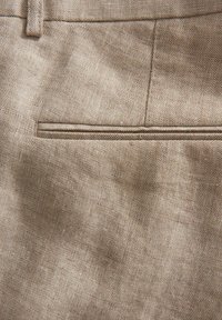 Tessuto di lino beige con una texture liscia, dotato di una tasca con profilo e un sottile motivo tessuto. La superficie presenta una leggera sfumatura di luce.
