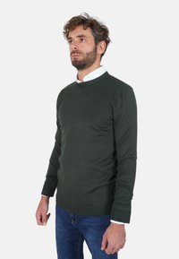 Maglione verde scuro lavorato a maglia con scollo tondo e polsini a coste, indossato sopra una camicia bianca con colletto, abbinato a jeans blu.