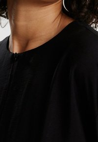 Haut noir avec un col rond, une fermeture à boutons sur la poitrine, et un tissu doux et texturé qui tombe harmonieusement.