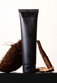 Tube noir à pression étiqueté « CRÈME POUR BOUCLES », 150 mL. Texte blanc décrivant l'utilisation du produit, sur fond de bois foncé et de noix de coco.