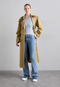 Beige trenchcoat med oversized passform, blå ribbestrikket topp, lyseblå jeans og blå sneakers. Viktige detaljer inkluderer store knapper og skuldre.