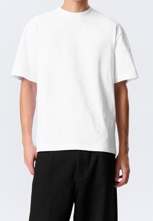 Homme portant un t-shirt blanc uni surdimensionné à manches courtes et un pantalon noir, debout devant un fond clair uni.