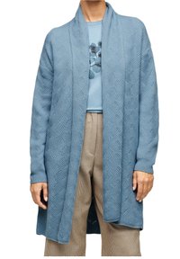 Cardigan azzurro chiaro realizzato in tessuto knit testurizzato con motivo a onde, aperto davanti e con maniche lunghe, indossato sopra un top azzurro chiaro e pantaloni a scacchi.