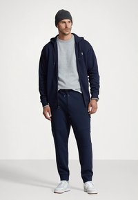 Sweatshirt azul-marinho com capuz e fecho de correr, t-shirt cinza de gola redonda, calças de fato de treino azul-marinho a condizer e gorro cinza de malha. O look fica completo com ténis com sola branca.