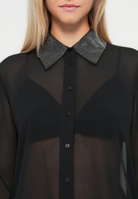 Bluză neagră din material transparent, cu un guler contrastant, embellizat. Gulerul este decorat cu mici strasuri strălucitoare. Butoanele din față sunt negre.