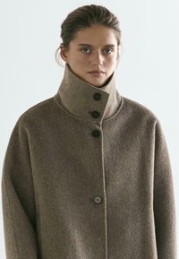 Brązowy wełniany płaszcz z wysokim kołnierzem, z trzema ciemnymi guzikami i luźną sylwetką, prezentowany na neutralnym tle.