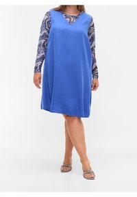 Blå bomullsklänning med ärmlös design, V-ringning och en mjuk textur, lager på lager över en långärmad mönstrad topp. Accentuerad med sandaler.