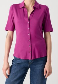 Fuchsia blouse met korte mouwen, een kraag en knoopsluiting, gemaakt van gladde stof met een subtiele glans; gecombineerd met blauwe denimbroek.