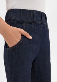 Ciemnoniebieskie dżinsowe spodnie z elastycznym pasem, z bocznymi kieszeniami i widocznymi detalami szwów. Gładka tekstura i dopasowany krój.
