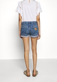 Jeansshorts med uppvikta fållar, två bakfickor med en zigzagdesign och en brun lädertag. Modellen bär en vit blus och sandaler.