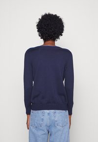 Personne aux cheveux noirs bouclés portant un pull à manches longues bleu marine et un jean bleu clair, debout contre un mur blanc uni.