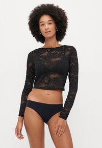 Czarny koronkowy crop top z długim rękawem, w kwiatowy wzór i z koralikowymi akcentami, zestawiony z czarnymi majtkami bikini. Modelka pozująca z neutralnym wyrazem twarzy.