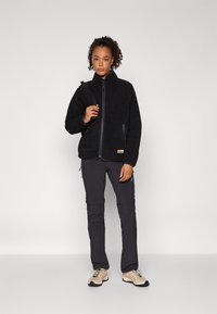 Giacca in fleece nera con colletto alto, chiusura a zip e tasche laterali. Indossata con pantaloni scuri e scarpe da trekking chiare.
