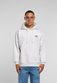 Helle graue Kapuzenjacke mit einer Fronttasche, verstellbarem Kordelzug und einem kleinen Logopatch. Tragen mit blauen Jeans.