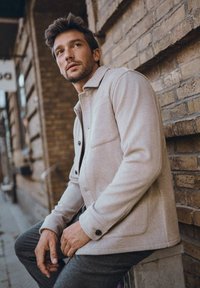 Ljus beige jacka i ullblandning med formell krage, två framfickor och svarta knappen detaljer, tillsammans med mörkgrå byxor.