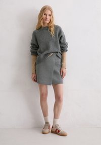 Grauer Strickpullover mit kastiger Form und hochgekrempelten Ärmeln, kombiniert mit einem passenden grauen Wickelrock, beigefarbenen Sneakern und weißen Socken.