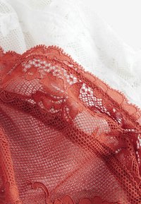 Laceweefsel in roest en wit. De roestkleurige kant heeft bloemmotieven met golvende randen, die delicate texturen en ingewikkelde details accentueren.
