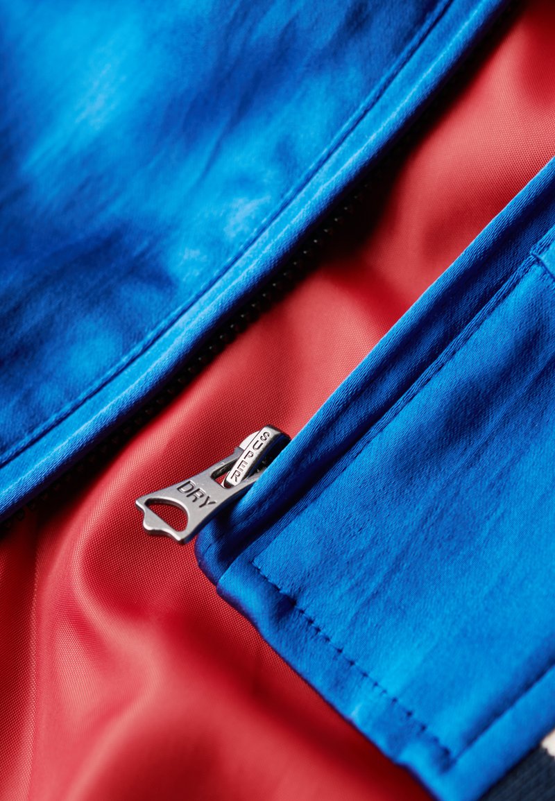 Tissu bleu avec une texture lisse et une doublure rouge. Comprend une fermeture éclair en argent marquée "DRY" et un détail de couture soigneusement fini.