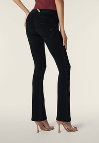 Nainen pukeutuneena mustiin istuviin bootcut-farkkuihin, joissa on takataskut, ja nudenvärisiin korkokenkiin, seisoo toisella kädellä lantiolla.