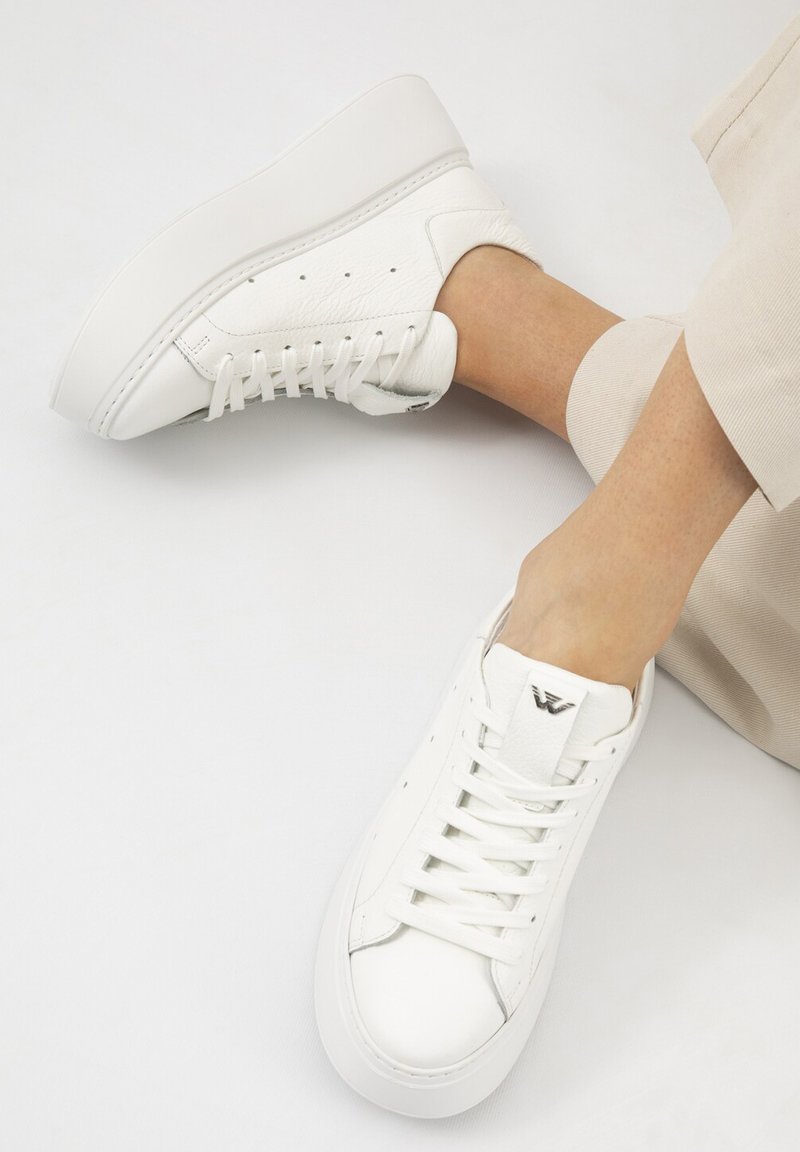 Witte leren platformsneakers met veters, gedragen door een persoon met een lichte huid en beige broek opgerold bij de enkel.