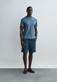 Fiatal férfi áll, kék rövid ujjú inget, mintás kék rövidnadrágot és sötétbarna mokaszint visel, egyszerű világos háttér előtt.