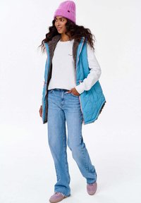 Akvově modrá prošívaná vesta se šedým fleecem uvnitř, nošená přes bílou halenku s dlouhým rukávem a světle modré džíny. Fialová pletená čepice a tenisky.