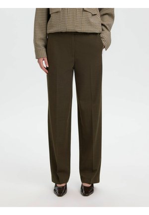 Femme portant un pantalon marron à jambes larges, une veste à carreaux et des chaussures pointues foncées, debout devant un fond clair uni.